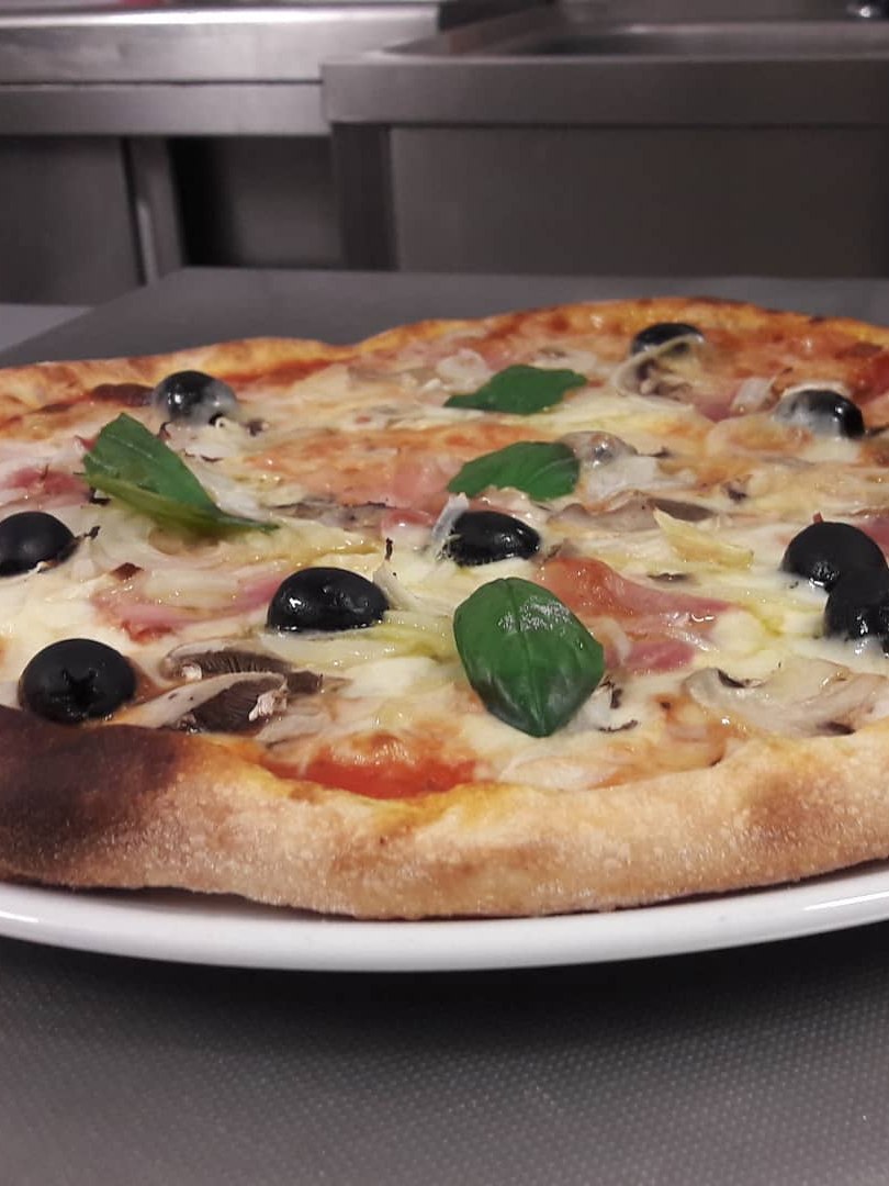 Pizza di Ristopizza da Tony con prosciutto, funghi, olive nere e basilico su piatto bianco in cucina d'acciaio
